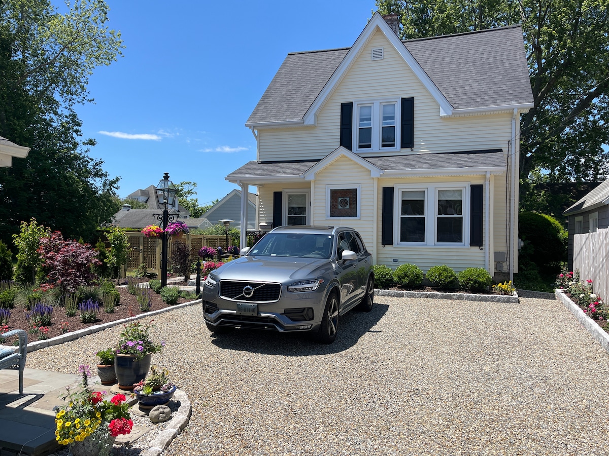 A charming yellow house is shown with a sloped roof and black shutters. A gray SUV is parked in the gravel driveway. Colorful flower beds surround the entrance, contributing to the welcoming exterior. The landscaping includes neatly trimmed shrubs and ornamental plants.
