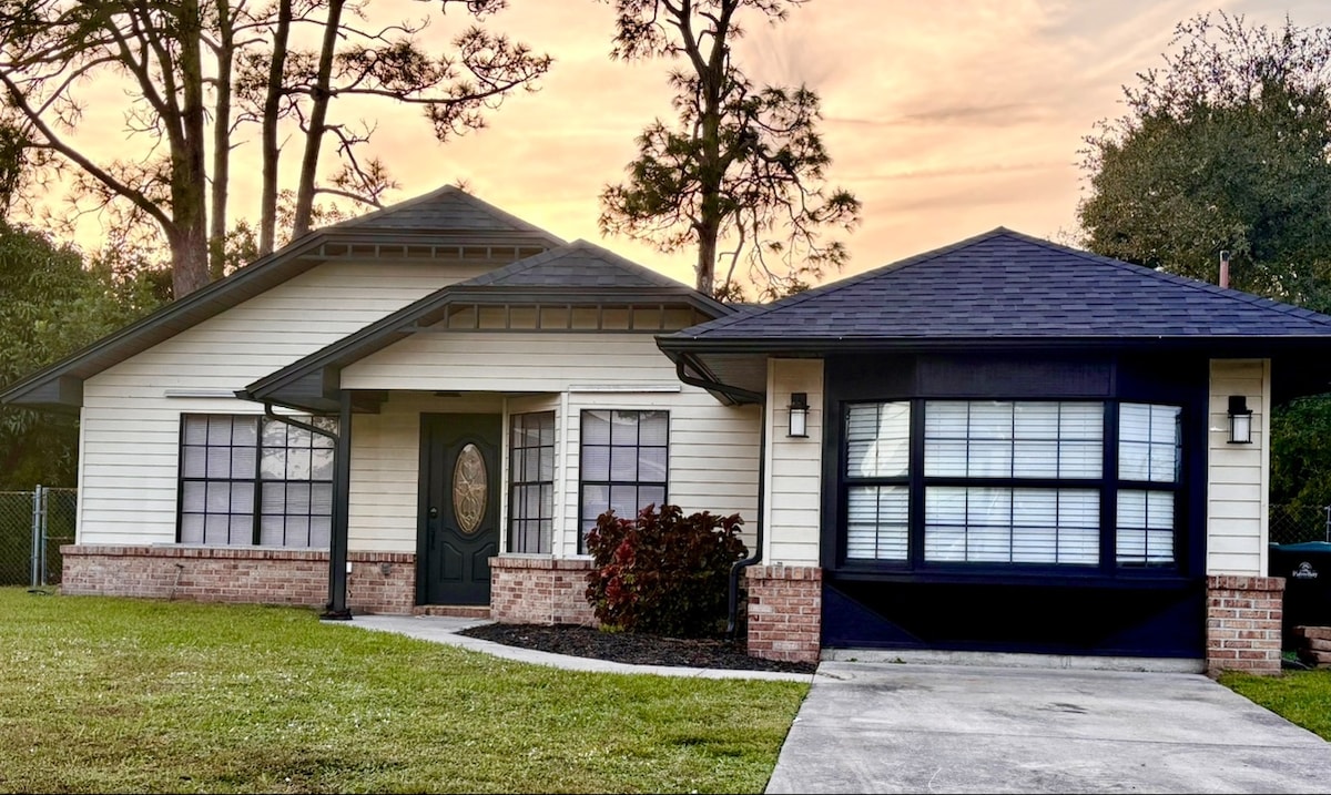 The exterior of a newly renovated home is depicted, featuring a yellow and black color scheme. A well-maintained lawn is present in front, with a walkway leading to the entrance. Large windows allow natural light to fill the interior, surrounded by greenery and trees.