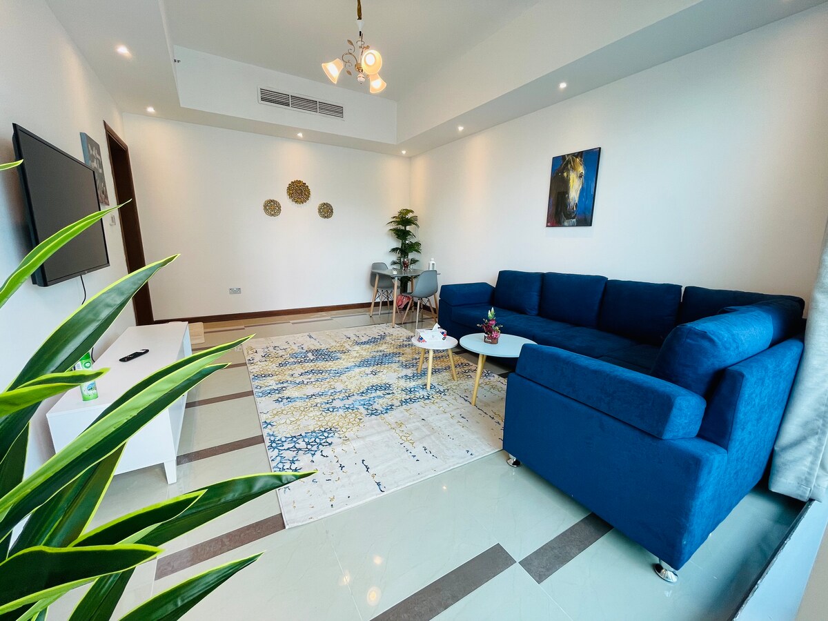 A modern living area is presented with a blue sectional sofa and a coffee table. A flat-screen television is mounted on the wall. Light decoratives are displayed, and a plant is positioned in the corner. A patterned rug adds texture to the tiled floor.