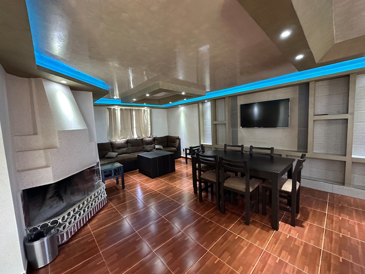 A spacious living area features a comfortable sectional sofa arranged around a central coffee table. A large dining table with seating for six is positioned nearby. Soft lighting and a sleek fireplace enhance the ambiance, while a wall-mounted television is visible.