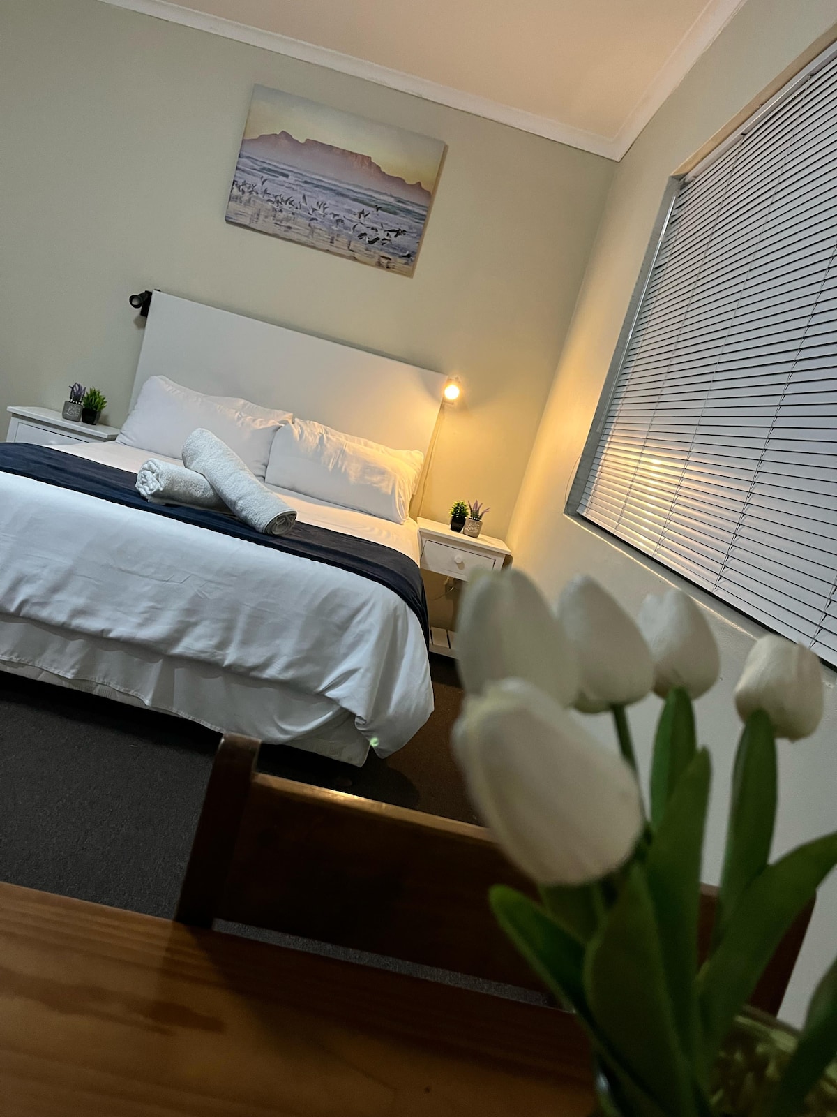 A cozy bedroom features a spacious bed with crisp white linens and a navy throw. A bedside table with a lamp stands beside the bed. In the foreground, a vase holds white tulips, and a scenic artwork hangs above the bed. Natural light filters through a window with blinds.