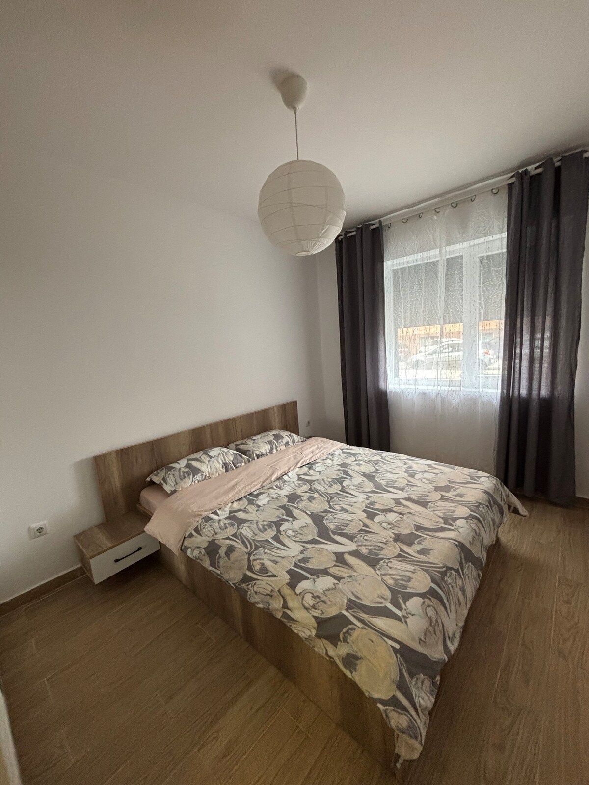 A cozy bedroom is presented with a spacious bed featuring a floral-patterned duvet. Natural light filters through a window covered by sheer and dark curtains. A simple nightstand is located beside the bed, enhancing the room's minimalistic design.