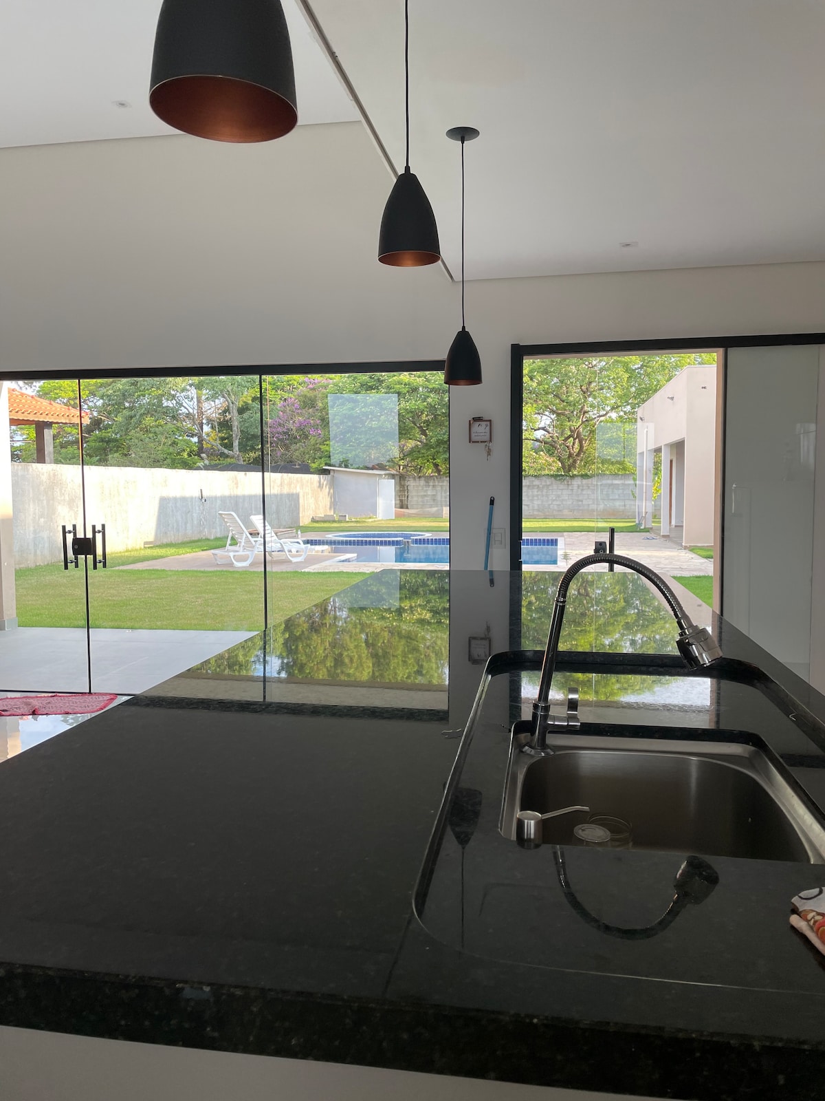 A modern kitchen space features a dark stone countertop with a stainless steel sink. Above, pendant lights provide subtle illumination. Large glass doors overlook a well-manicured yard with a pool, enhancing the connection between indoor and outdoor areas.