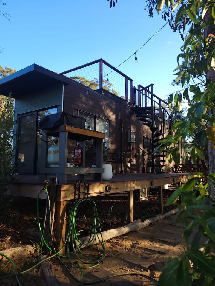 A modern cabin with a raised wooden deck and a spiral staircase leading to a rooftop patio. The deck features an outdoor grill and seating area, surrounded by trees and greenery. A garden hose is coiled underneath the deck, adding to the rustic outdoor setting.