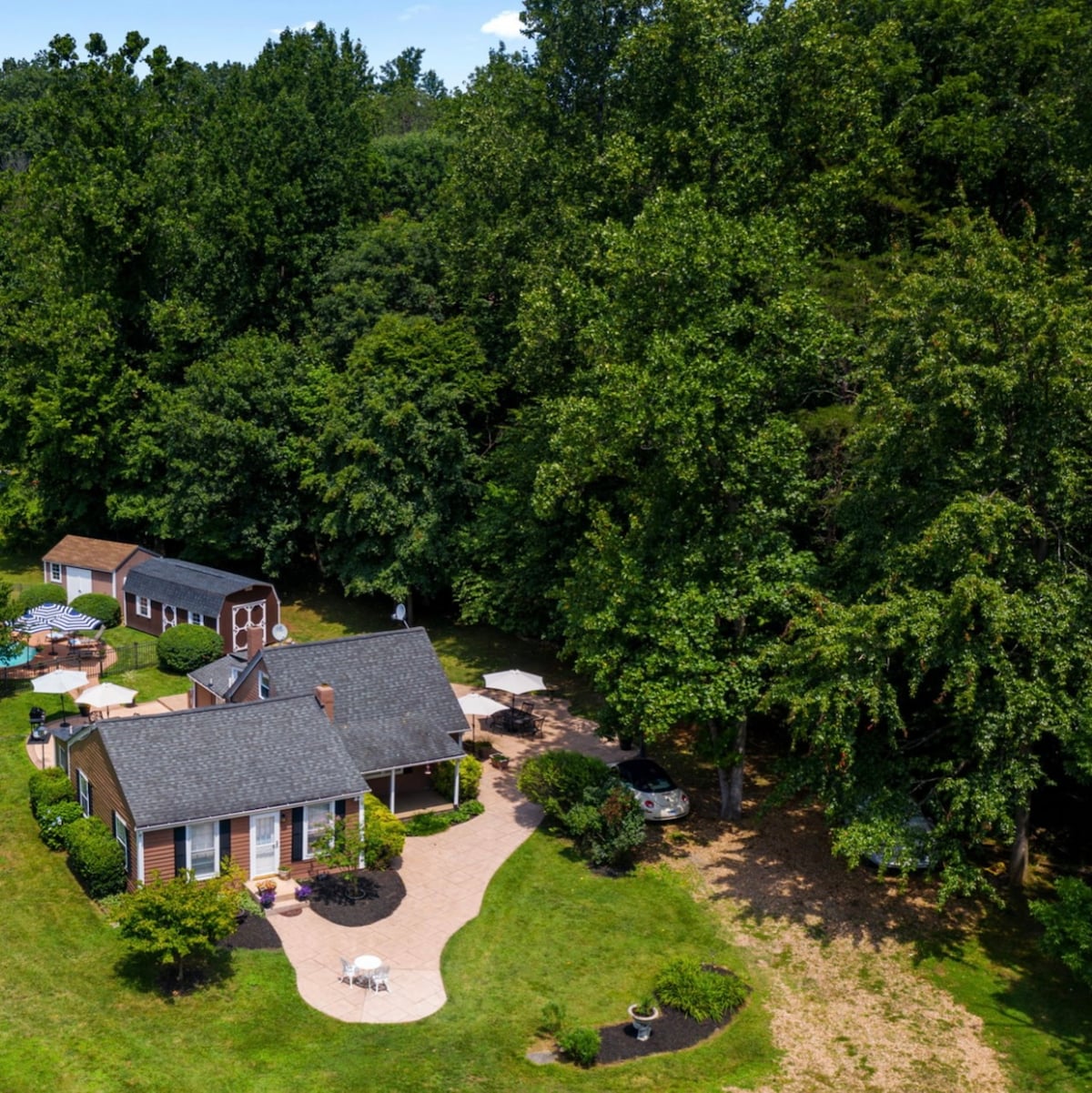 An aerial view of a charming cottage surrounded by lush greenery, showcasing a well-maintained lawn. A winding pathway leads to the house with a patio area, while a pool is visible in the background, enhancing the tranquil setting.