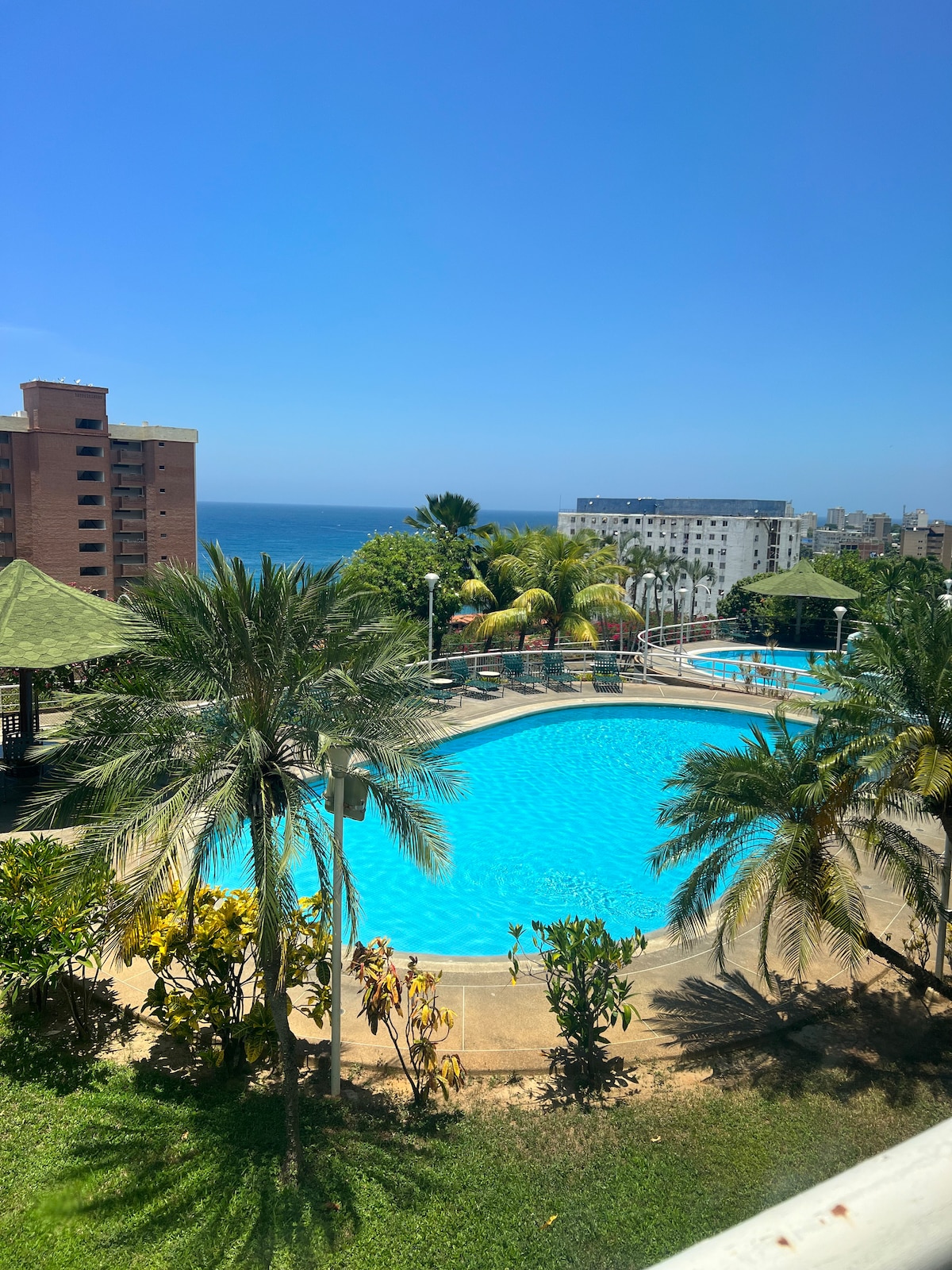 A serene outdoor area features two swimming pools surrounded by lush greenery. Palm trees and colorful foliage enhance the scenery, with a distant view of the ocean visible under a clear blue sky. A shaded gazebo provides additional outdoor relaxation space.