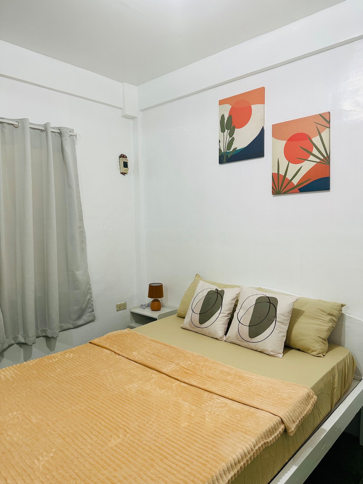 A light-filled bedroom features a comfortable bed adorned with neutral-colored linens and decorative pillows. Two pieces of wall art with natural motifs add subtle color above the bed. Sheer curtains gently filter the light, enhancing the space's calm ambiance.