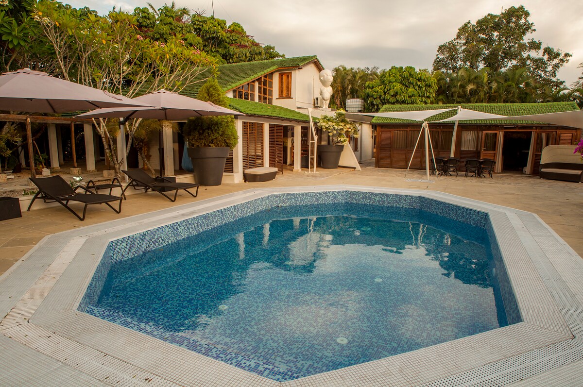 A spacious outdoor area features a crystal-clear octagonal pool surrounded by lounge chairs and shaded by large umbrellas. Lush greenery and well-maintained landscaping enhance the tranquil setting, with a few wooden structures visible in the background.