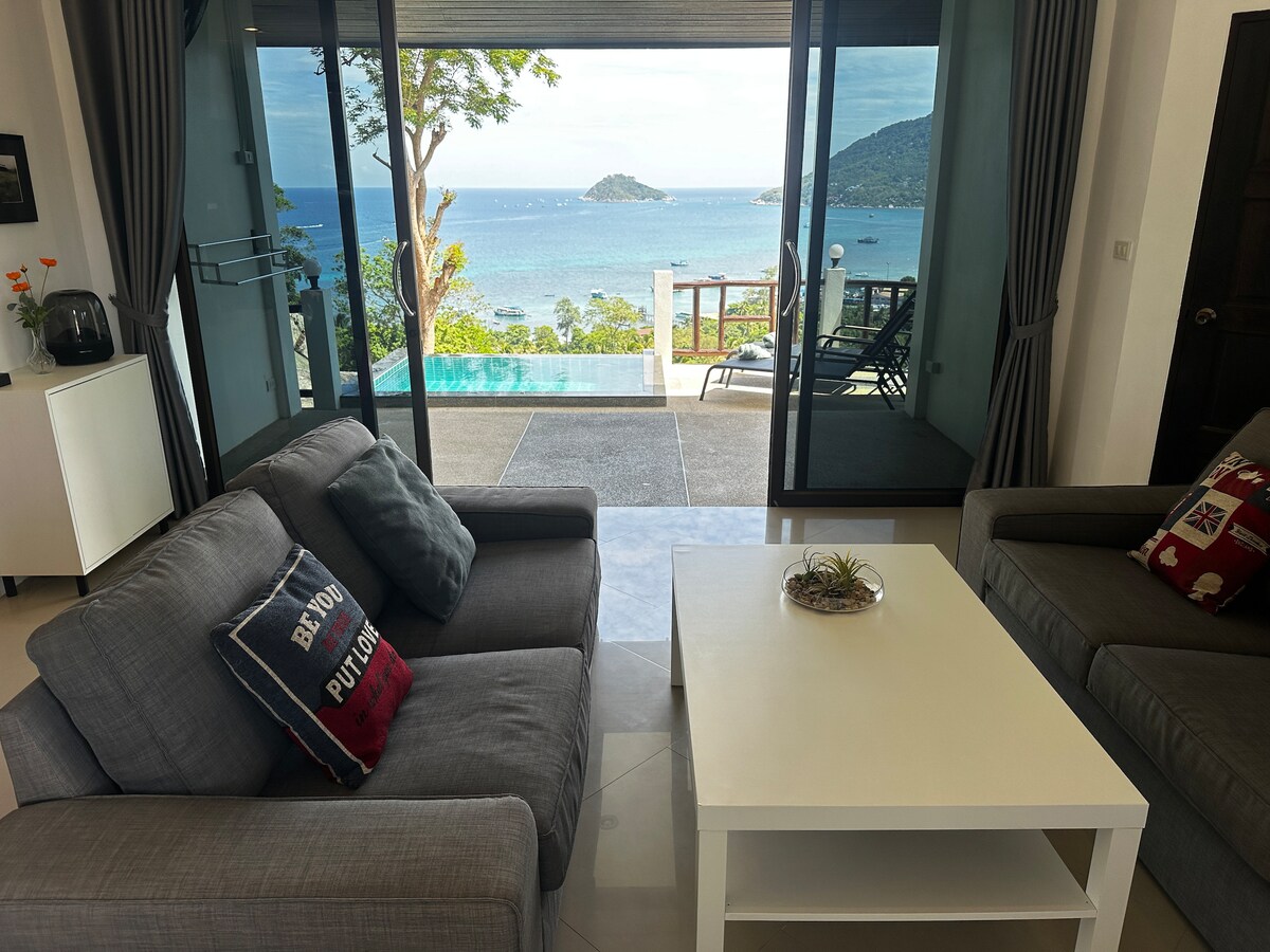 A spacious living area exhibits large glass doors that open to reveal a stunning ocean view. Comfortable sofas are arranged around a coffee table. A glimpse of the private plunge pool can be seen in the background, framed by lush greenery.