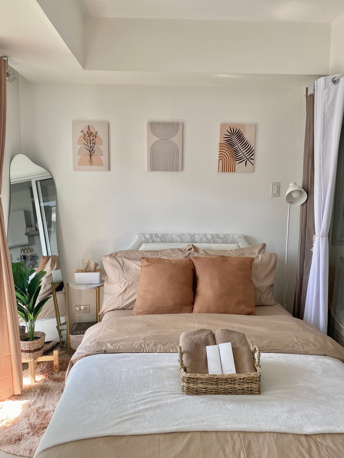 A serene bedroom is presented, featuring a spacious bed adorned with plush pillows. Artwork in soft tones hangs above the bed. A full-length mirror reflects natural light, while a plant adds a touch of greenery. A tray with rolled towels rests on the bed.
