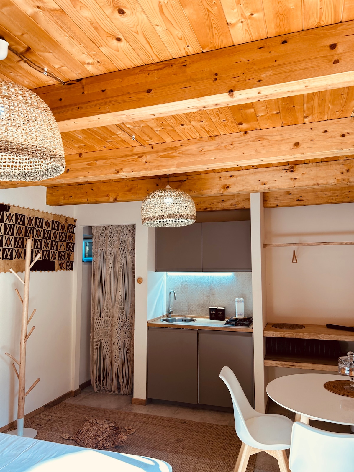 The interior features a wooden ceiling, complemented by woven light fixtures. A compact kitchenette with modern appliances is visible, alongside a dining area with a round table and white chairs. A cozy seating nook is positioned against the wall, enhancing the functional layout.