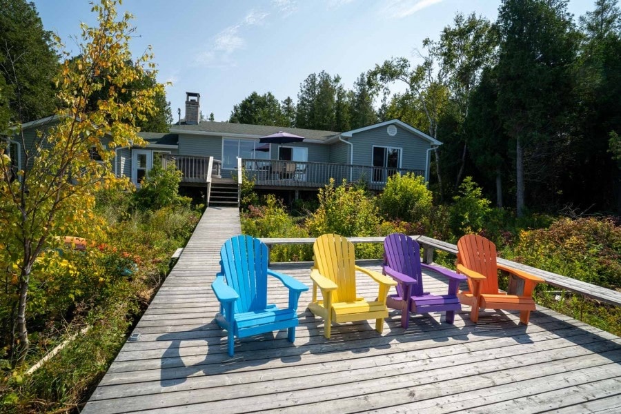 Four colorful adirondack chairs sit at the end of a wooden dock, facing the cottage and surrounding greenery. The cottage features a spacious deck with a purple umbrella and is framed by trees, creating a serene outdoor setting.