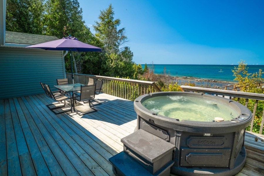 An outdoor deck overlooks the water, featuring a hot tub and a dining set shaded by an umbrella. The calm lake sparkles under clear skies, with lush greenery lining the shore, enhancing the tranquil setting.