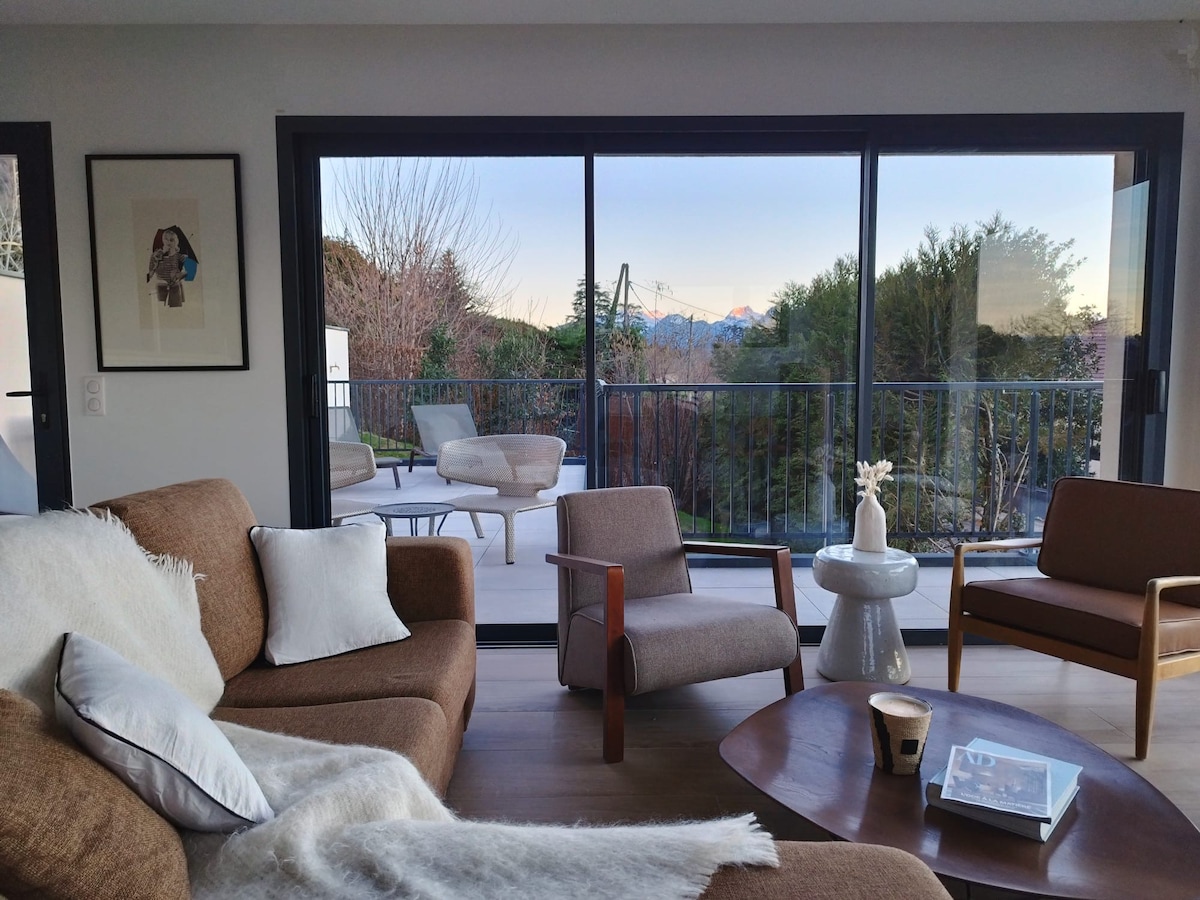 A modern living space features a comfortable brown sofa and two armchairs arranged around a wooden coffee table. Large glass doors provide a view of the outdoor area, framed by trees and mountains in the distance.