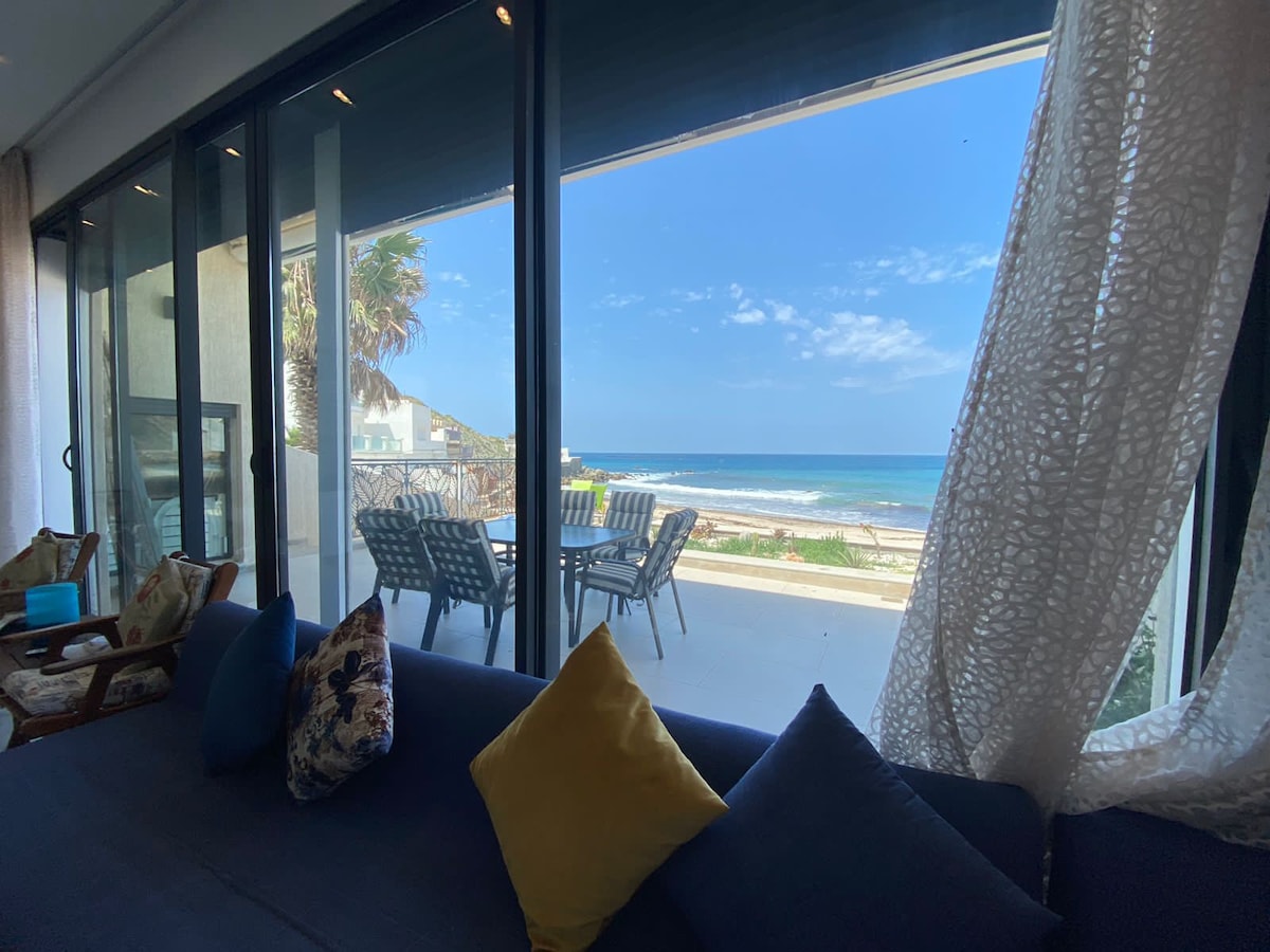 A spacious living area is featured with large glass doors that open to a private terrace overlooking the beach. Outdoor dining furniture is visible on the terrace, while bright blue cushions and textured throws adorn the sofa, enhancing the view of the ocean.
