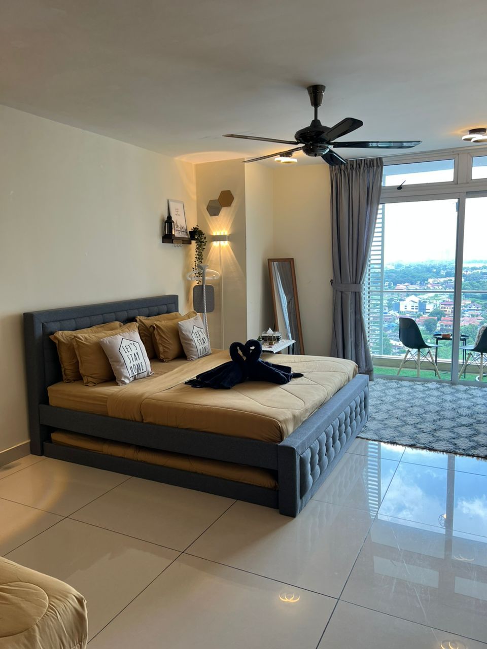 A modern bedroom features a spacious bed adorned with neutral-toned linens and decorative pillows. A large window allows natural light to fill the room, while a balcony with seating is visible in the background. A ceiling fan adds a touch of comfort to the space.