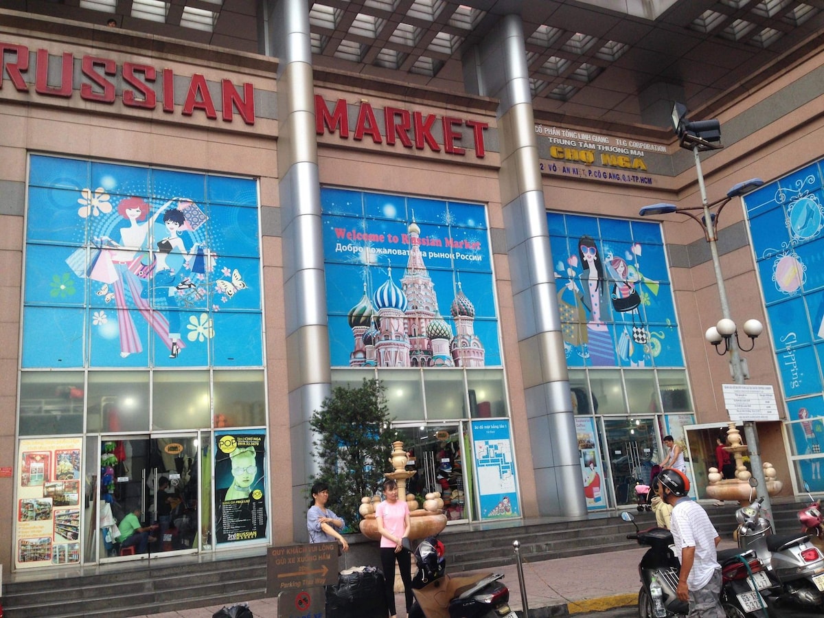 The exterior of the Russian Market features large glass windows adorned with vibrant murals depicting cultural scenes. The market entrance is accessible, with people present and scooter parking visible in the foreground. Bold red signage indicates the market name, reflecting local architecture.