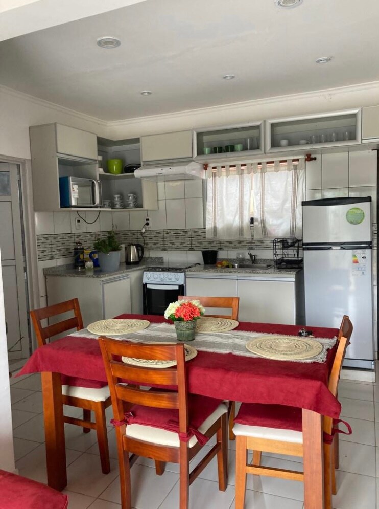 The kitchen area features modern cabinetry in soft gray tones and a spacious countertop. A dining table is set with four wooden chairs and decorative placemats. Light filters through a sheer curtain, illuminating the room, which includes essential appliances such as a refrigerator and stove.