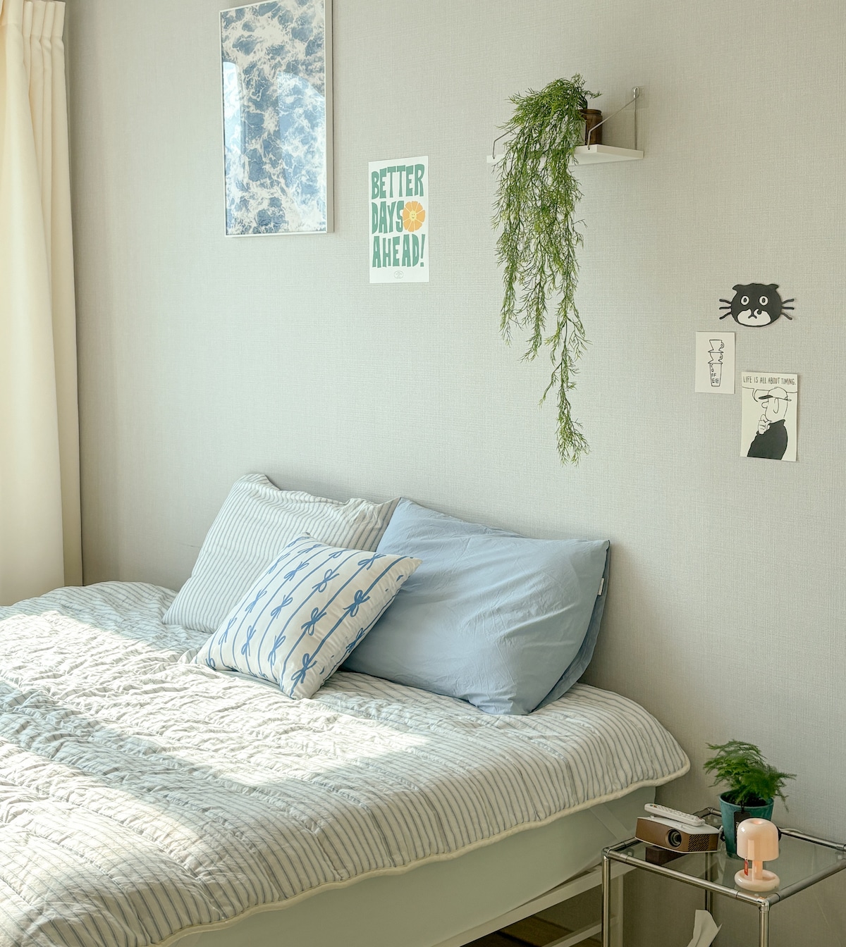A peaceful bedroom features a spacious bed with light blue and striped bedding. Green wall plants and framed prints are visible on the light-colored walls. Soft natural light enters through the sheer curtains, enhancing the calming atmosphere.