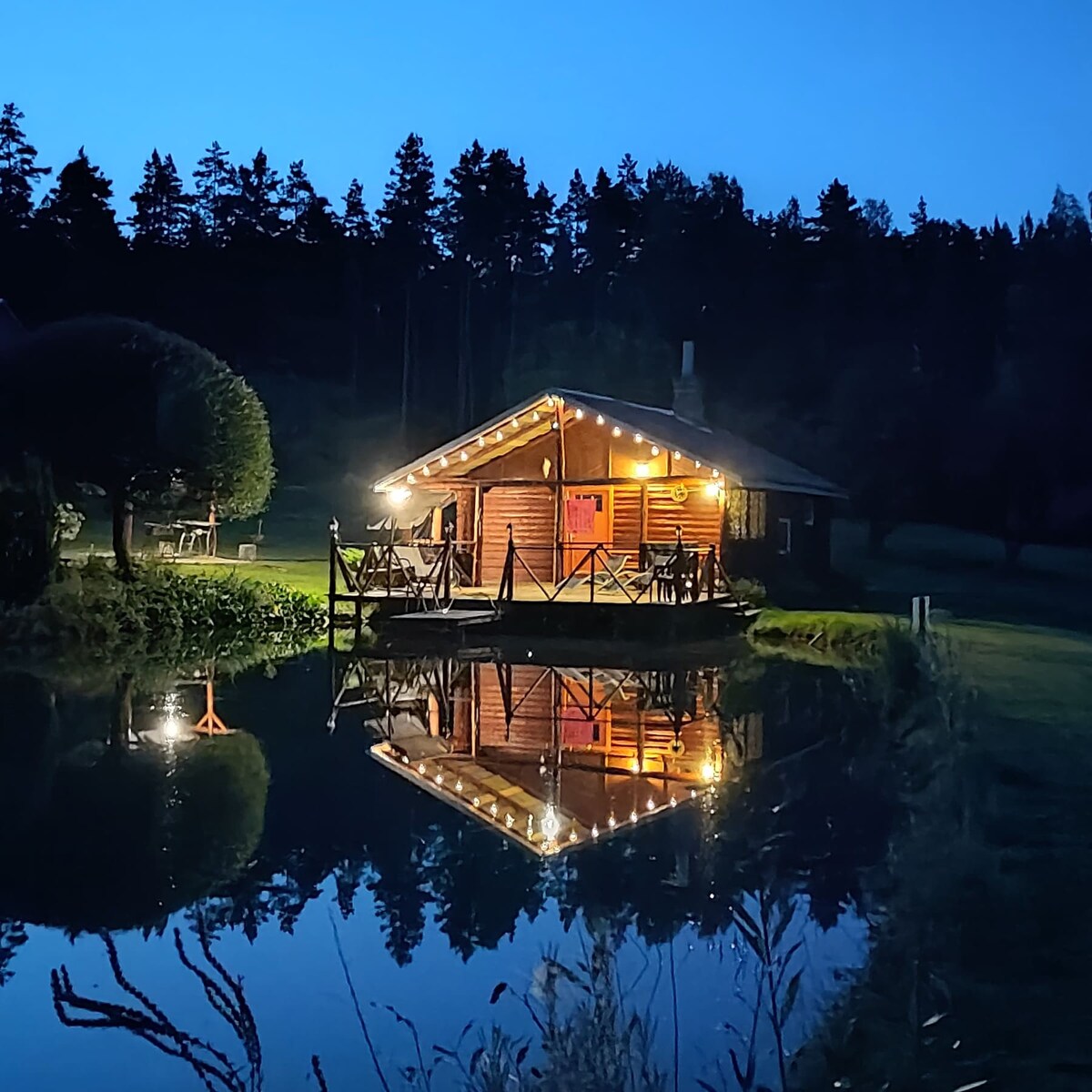A charming log cabin is set beside a serene pond, reflecting the warm glow of outdoor lights. Surrounding trees create a natural backdrop, while a wooden deck extends over the water, offering a peaceful retreat. The scene conveys tranquility, ideal for family relaxation.