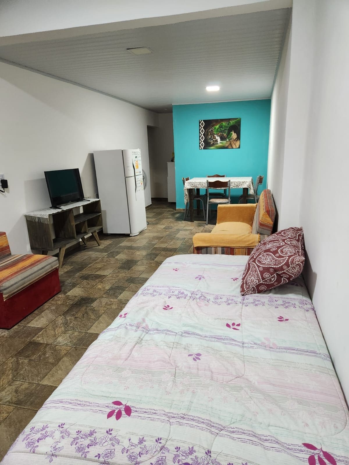 An inviting living area features a spacious daybed with a patterned cover, complemented by a colorful armchair and a table set for dining. A television and refrigerator are positioned against a neutral wall, with a vibrant turquoise accent wall enhancing the space.