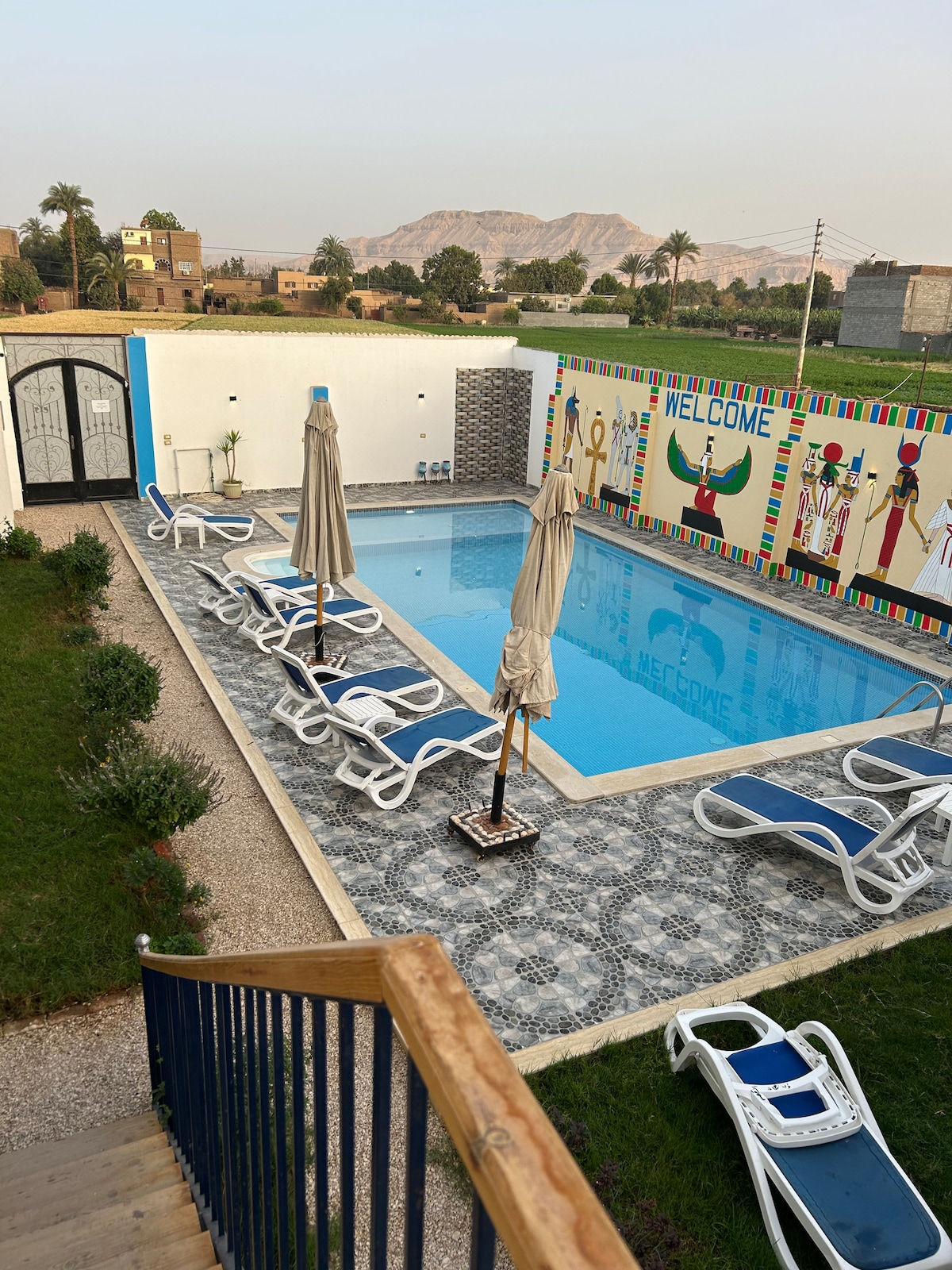 A serene pool area is surrounded by lounge chairs and umbrellas, providing a relaxing space. Colorful artwork featuring welcoming designs is displayed on the adjacent wall. Lush greenery and distant mountains complete the tranquil setting, inviting guests to unwind by the water.