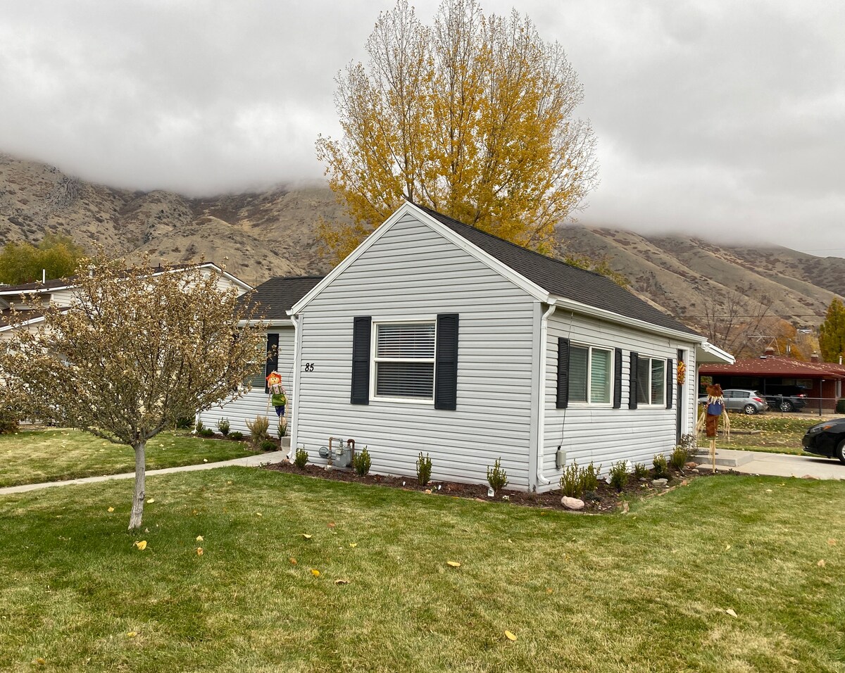 A charming cottage is set against a backdrop of mountains, with a well-maintained lawn and trees. The exterior features light-colored siding, dark shutters, and a welcoming entrance. A small flowerbed and pathway frame the home, providing a peaceful atmosphere.