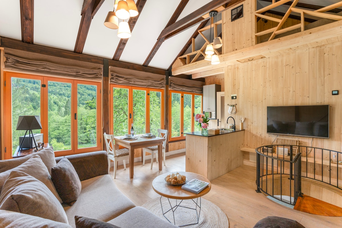 A spacious living area features large windows with orange frames, allowing natural light to fill the space. The open layout includes a kitchen with a breakfast bar and a dining table, while a cozy sofa invites relaxation. A round dining table is set for meals.