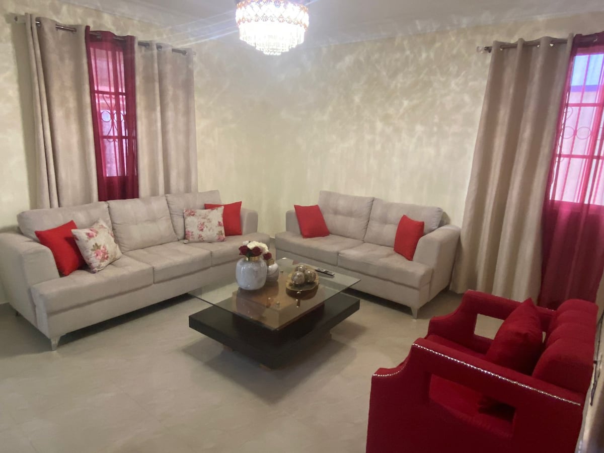 A bright living area is furnished with two light-colored sofas adorned with decorative pillows, a red accent chair, and a sleek glass coffee table. Natural light flows through the large windows, framed by soft curtains, enhancing the room's spacious feel.