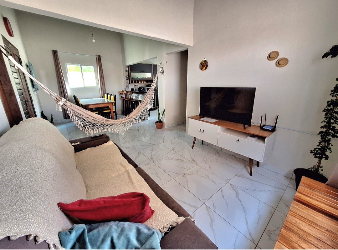 An open living area features a comfortable sofa with neutral tones and colorful cushions. A hammock hangs invitingly nearby. A TV sits on a modern cabinet while a dining area with a table is visible in the background, providing a cozy yet spacious atmosphere.