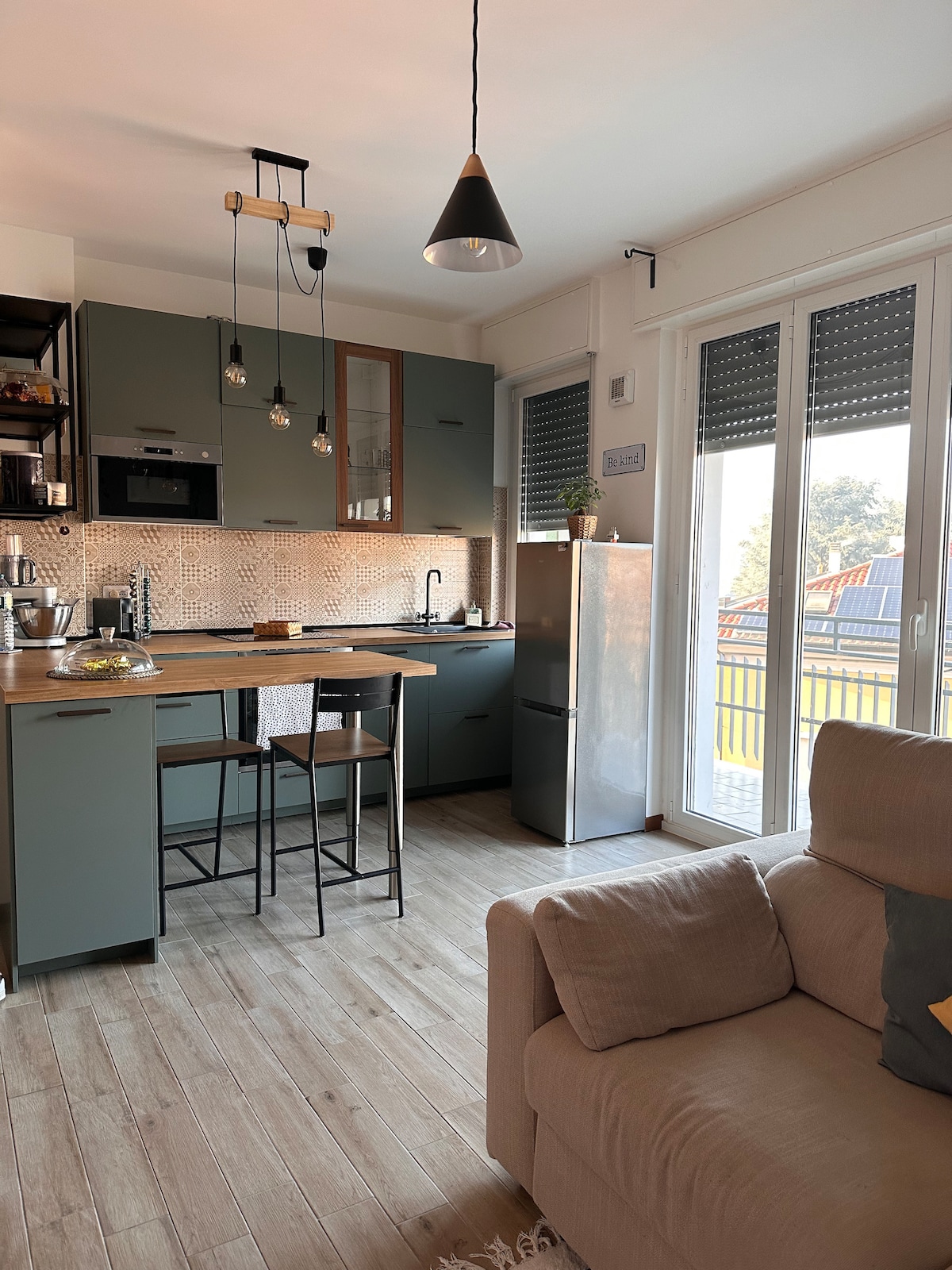 A modern kitchen area is equipped with sleek cabinetry and a countertop, paired with two high stools. Nearby, a stylish refrigerator and a cozy seating area are visible. Large windows allow ample natural light, with a view of the balcony and surrounding greenery.