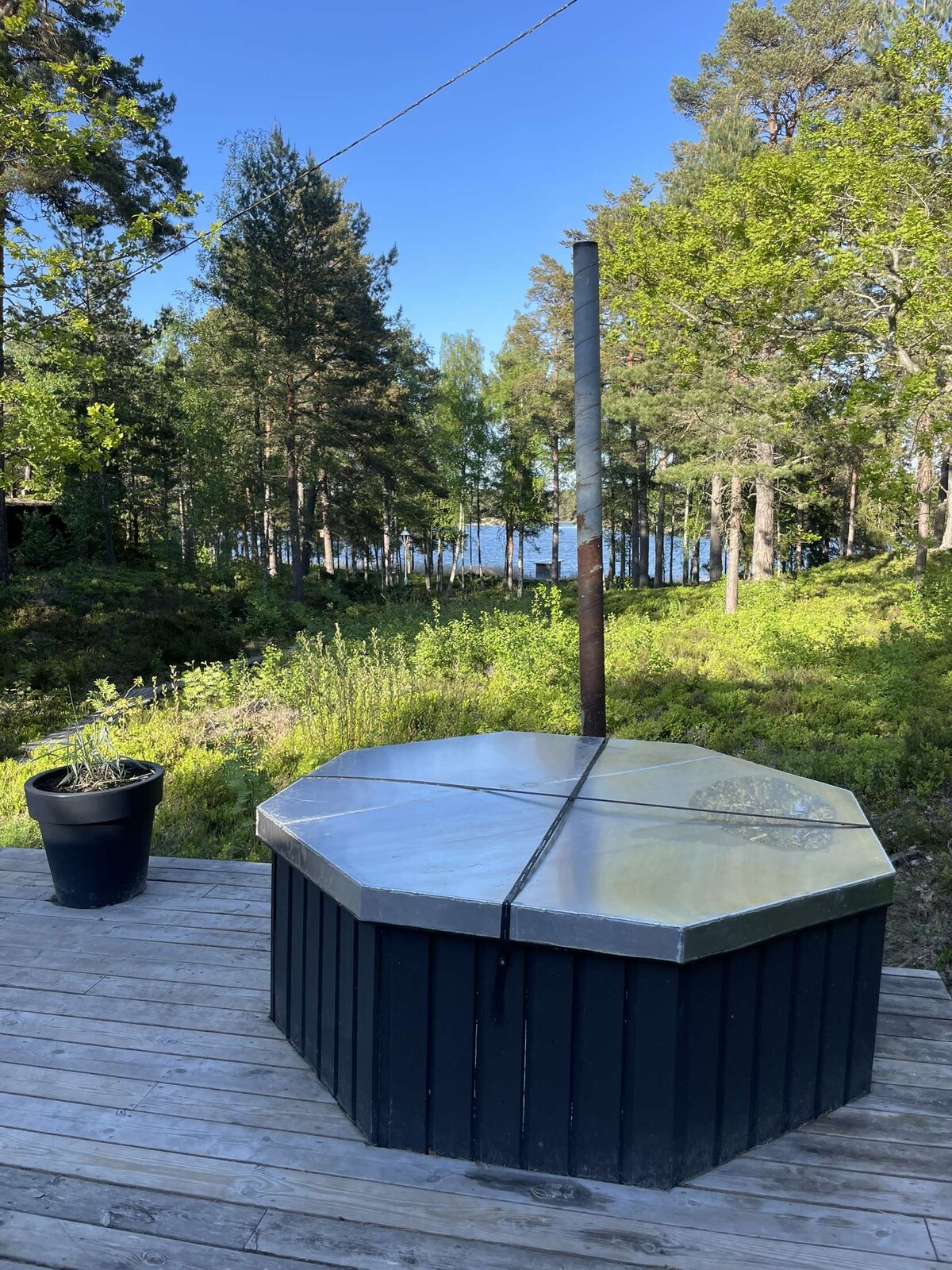 An outdoor hot tub is situated on a wooden deck, surrounded by lush greenery. The octagonal structure features a metal top and a chimney, with a view of the tranquil lake visible in the background through the trees.