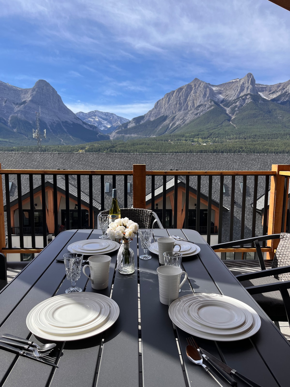 An outdoor dining table is set with plates and glasses, overlooking majestic mountain views. The scene features a fresh flower arrangement at the center, with a clear sky and several chairs positioned around the table, suggesting a comfortable space for meals amidst nature.
