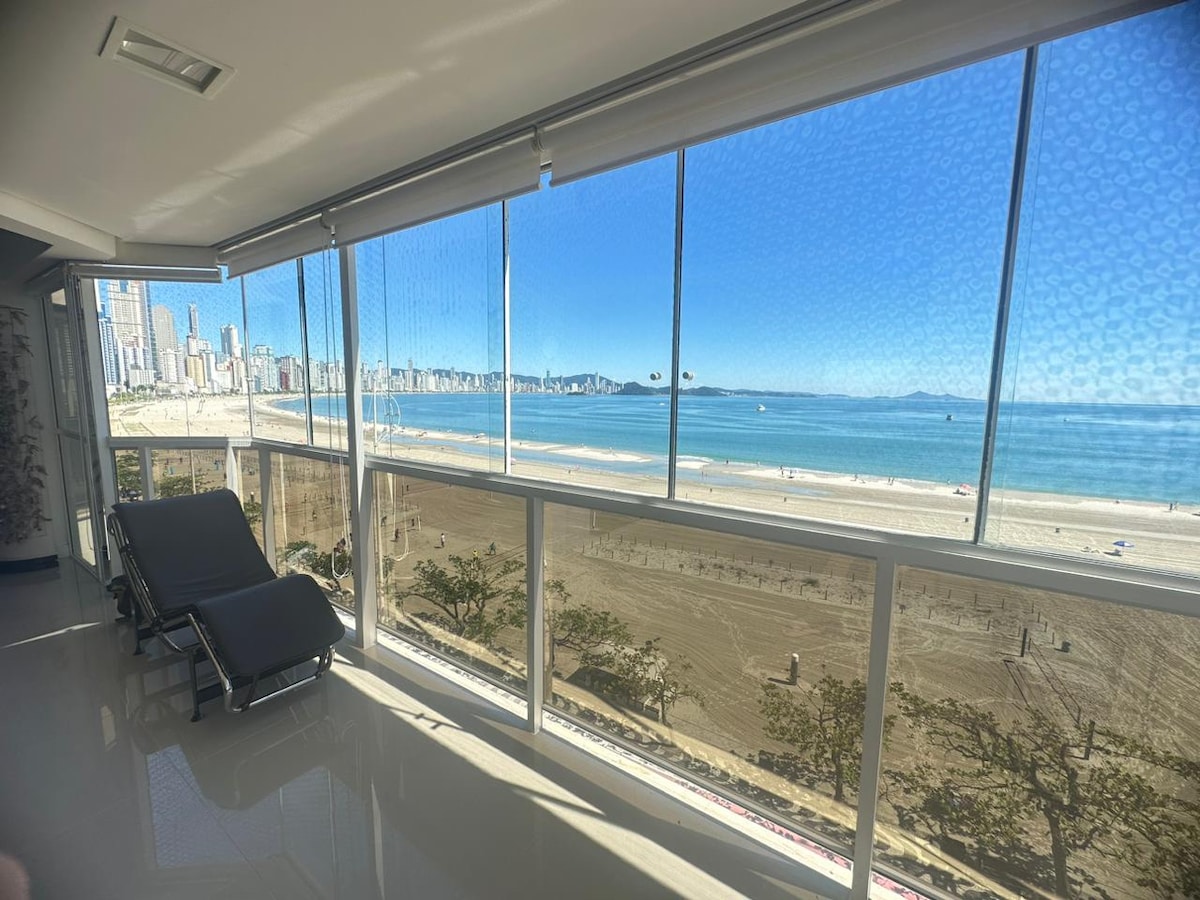 A bright and airy space features large windows offering stunning views of the beach and coastline. Two lounge chairs are positioned beside the window, inviting relaxation while enjoying the scenery. The smooth, polished floor reflects ample natural light, creating an open and serene atmosphere.