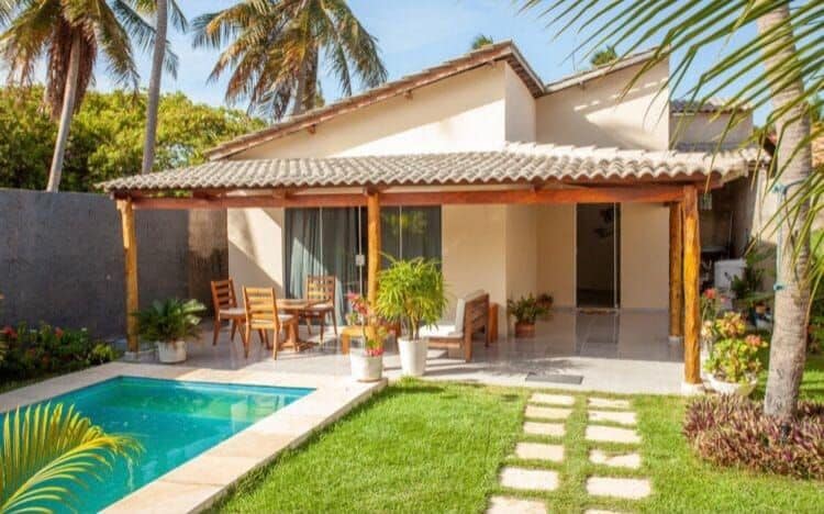 A tranquil outdoor space features a small swimming pool surrounded by lush greenery. Wooden chairs and a table are positioned on a covered patio, with a pathway of stones leading through the well-maintained garden.