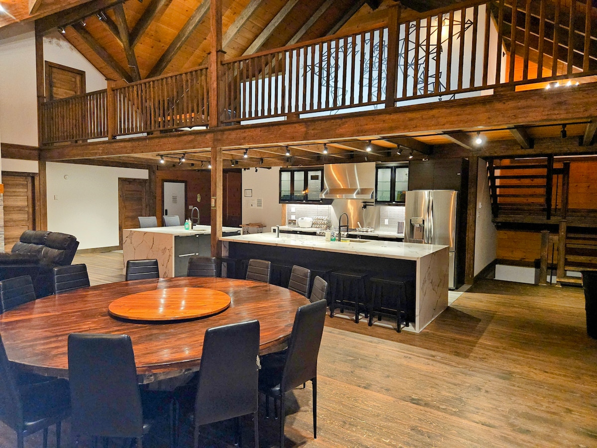 An open-concept living space features a large round dining table surrounded by black chairs. A modern kitchen with stainless steel appliances is visible, with ample counter space and cabinetry. High wooden ceilings with exposed beams contribute to the spacious atmosphere, along with a lofted area above.