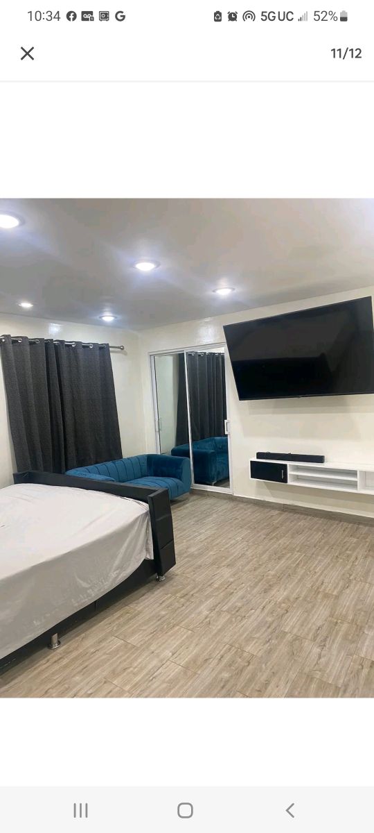 A modern sleeping area features a spacious bed covered with a light sheet. A blue sofa is positioned nearby, and a large flat-screen TV is mounted on the wall opposite the bed. Floor-to-ceiling curtains frame the mirror closet, complementing the room's neutral tones.