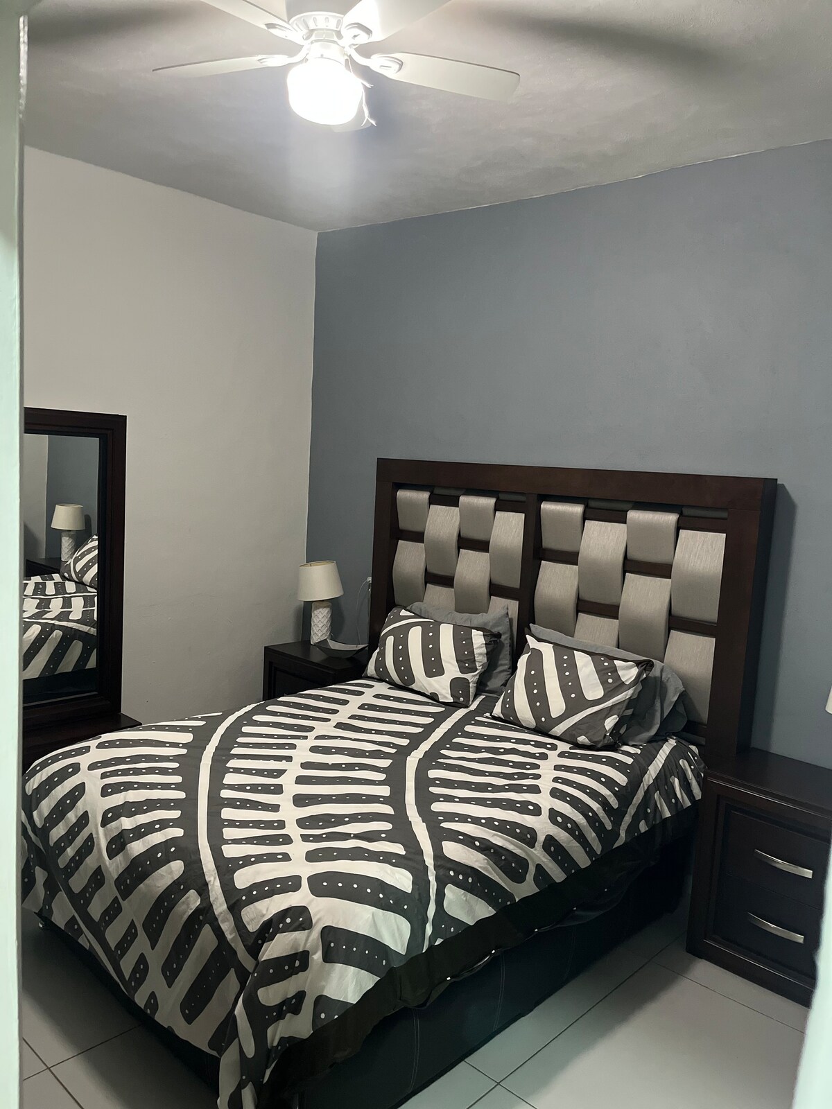 A comfortable bedroom features a spacious bed with a patterned duvet and two matching pillows. A wooden headboard complements the design. Nightstands with lamps are positioned on either side, and a full-length mirror reflects the room's layout.