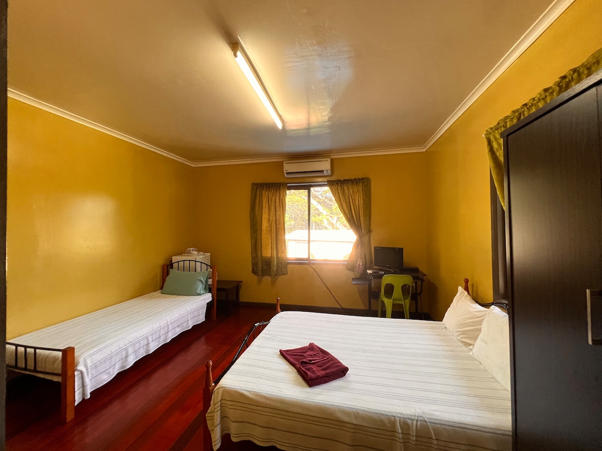 A well-lit room features a spacious bed and a single bed, both dressed in white linens. Yellow walls create a warm atmosphere, while windows with green curtains allow natural light to illuminate the space. A desk with a chair is positioned nearby.