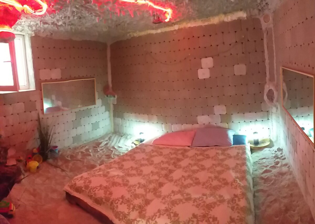A serene salt room features walls constructed from salt bricks, creating a unique atmosphere. The spacious bed is positioned at the center, with low bedding allowing for easy access to the salt-covered floor, highlighted by soft illuminated areas. Mirrors are present on opposite walls.