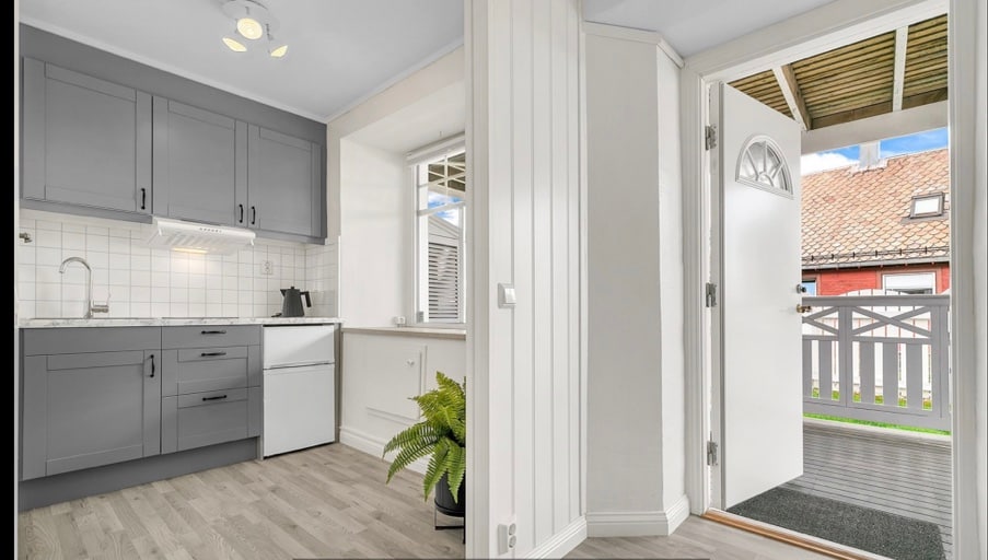 A compact kitchen features gray cabinetry, white countertops, and essential appliances including a refrigerator and stove. Adjacent, an entryway is highlighted by a door leading to an outdoor area, with a small potted plant adding a touch of greenery.