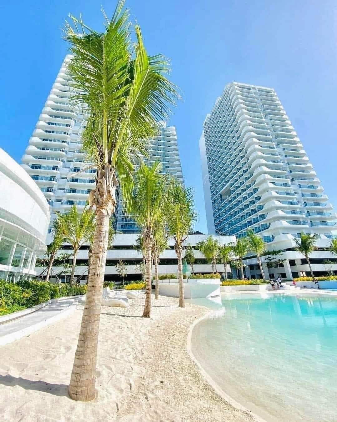 Two modern high-rise buildings are viewed from a sandy beach area, surrounded by palm trees. A serene pool reflects the clear blue sky, while landscaped greenery enhances the inviting outdoor space.