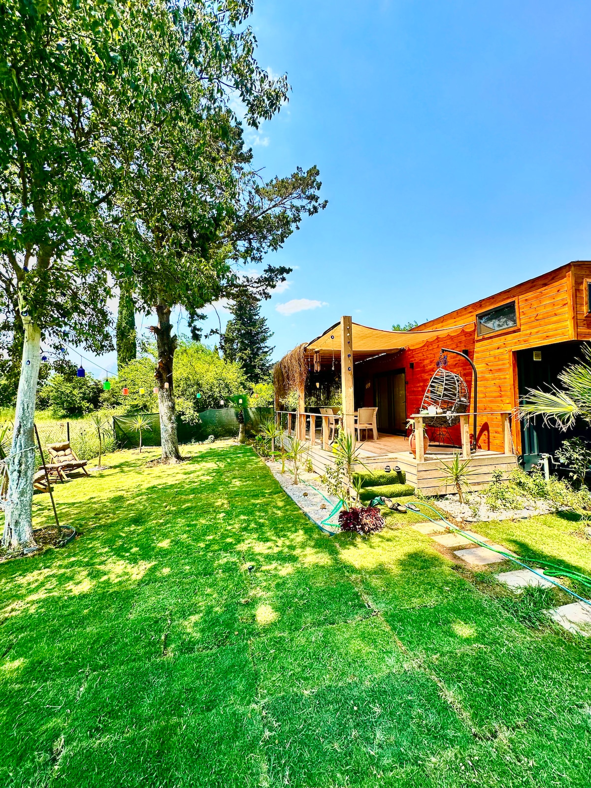 A sunny outdoor area features well-maintained grass and various plants, enhancing the natural ambiance. The wooden house is partially visible, showcasing a deck area with modern outdoor seating. Trees provide shade, contributing to a peaceful outdoor environment.