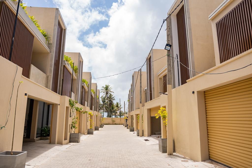A well-maintained courtyard is shown, flanked by modern two-story buildings. Each building features large balconies with wooden slats and potted plants. The wide pathway leads toward palm trees in the background, brightened by natural light and partially cloudy skies.