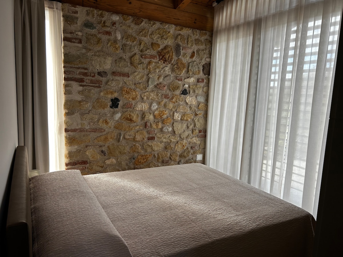 A spacious bed is positioned against a textured stone wall, illuminated by natural light filtering through sheer curtains. The warm tones of the stone contrast with the soft fabric of the bedding, creating a serene sleeping environment.