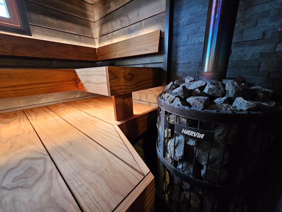 The image showcases a wooden sauna interior with smooth wood paneling and benches. A stone sauna stove is visible with a metallic finish and a basket of stones. Warm lighting highlights the natural textures, creating a serene atmosphere.