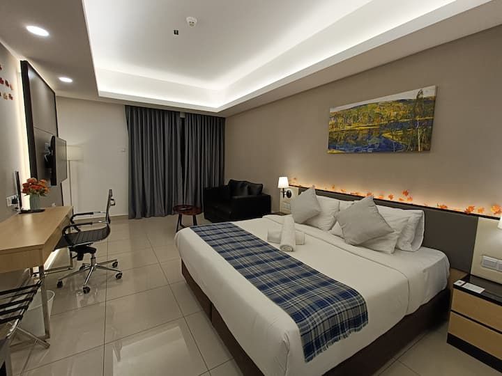 A spacious room is furnished with a king bed topped with decorative pillows and a cozy throw. A study desk and a chair are present, alongside a small seating area with a dark sofa. Natural light floods the room through floor-to-ceiling curtains.