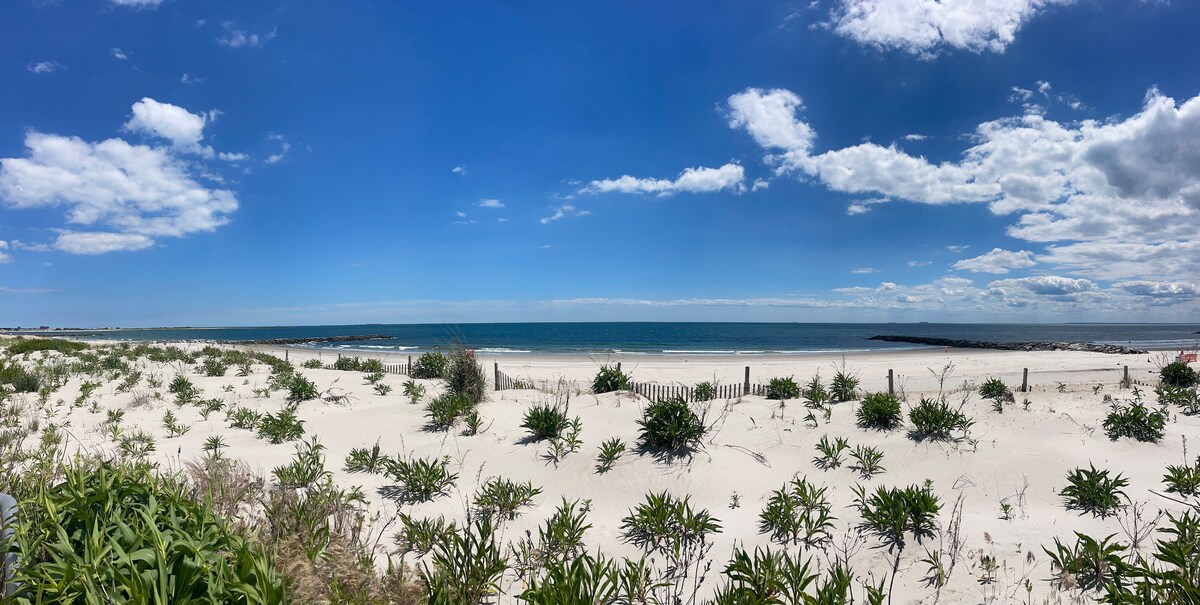 A panoramic view of a sandy beach is presented, featuring gentle dunes covered with low greenery. The calm ocean meets a clear blue sky with scattered clouds, creating a serene coastal landscape. A distant jetty is visible at the horizon, enhancing the scene's tranquil atmosphere.