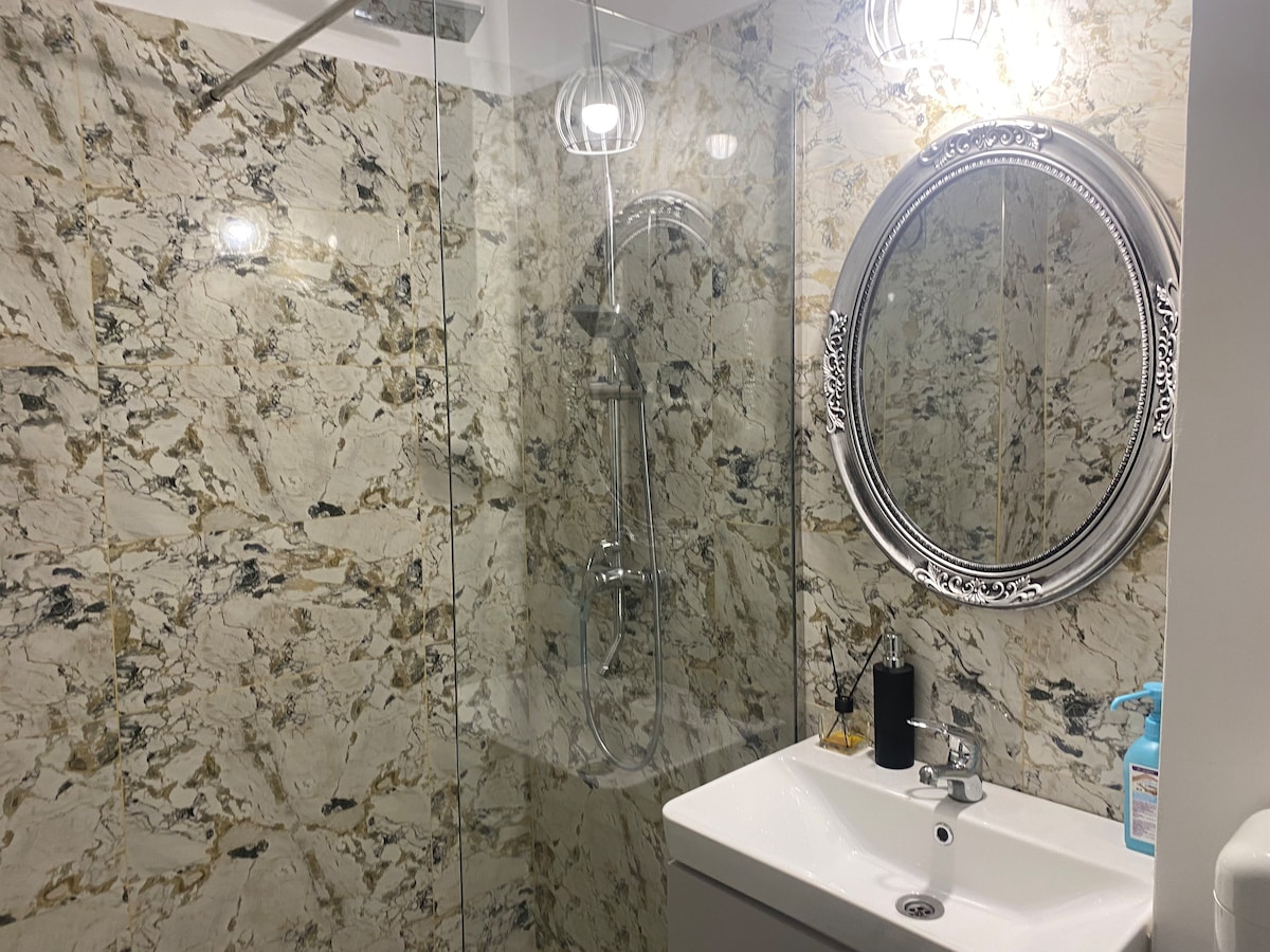 The bathroom features a modern design with marble-patterned walls. A spacious glass shower enclosure is visible, complemented by a sleek round mirror above a white sink. Soft lighting from fixtures adds a gentle glow to the space.