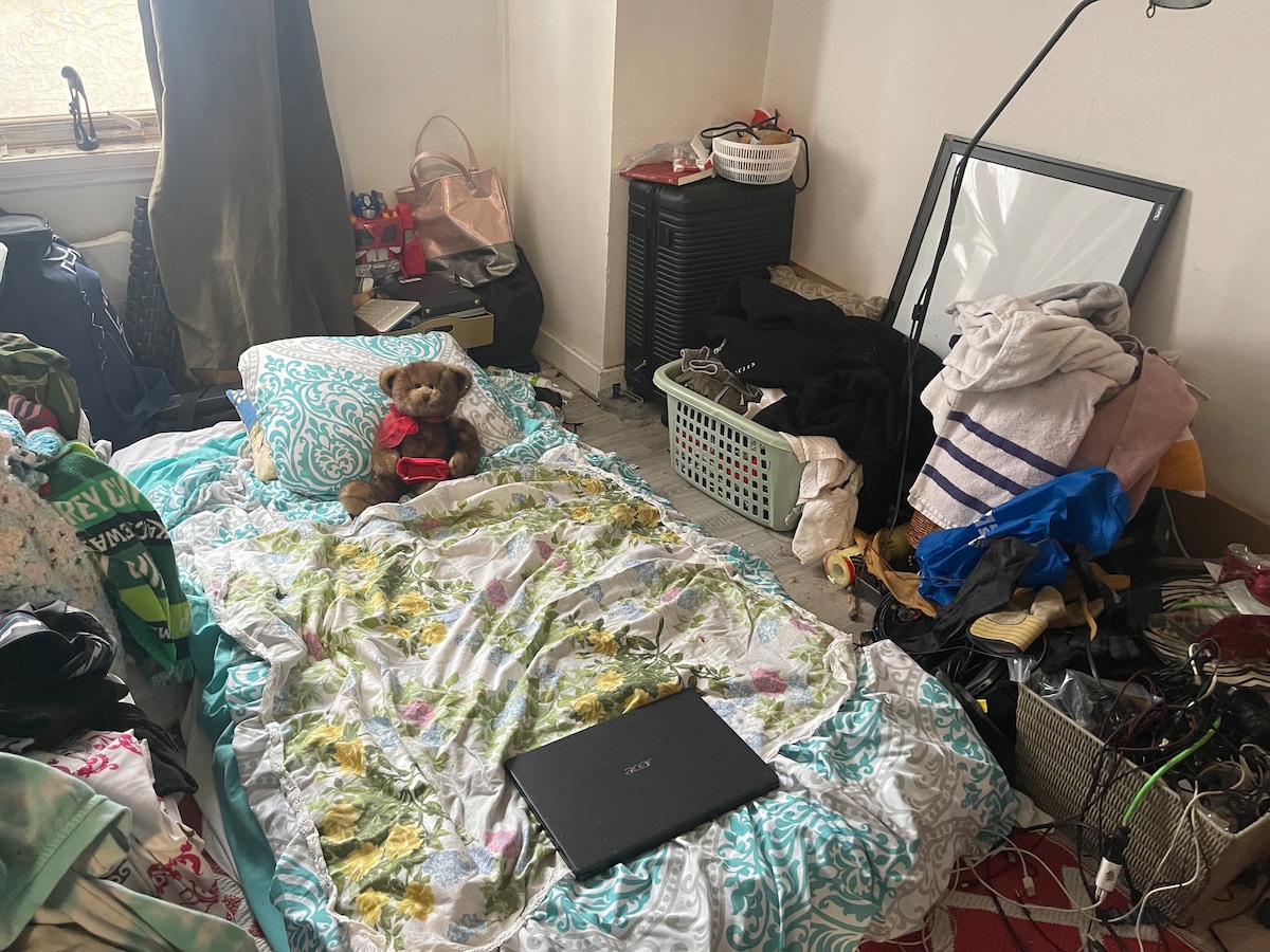 A sleeping area is presented with a floral-patterned comforter laid over a spacious bed on the floor. A laptop rests nearby, and a teddy bear is positioned on the bed. Various bags and clothing items are scattered around, alongside laundry baskets and a mirror.